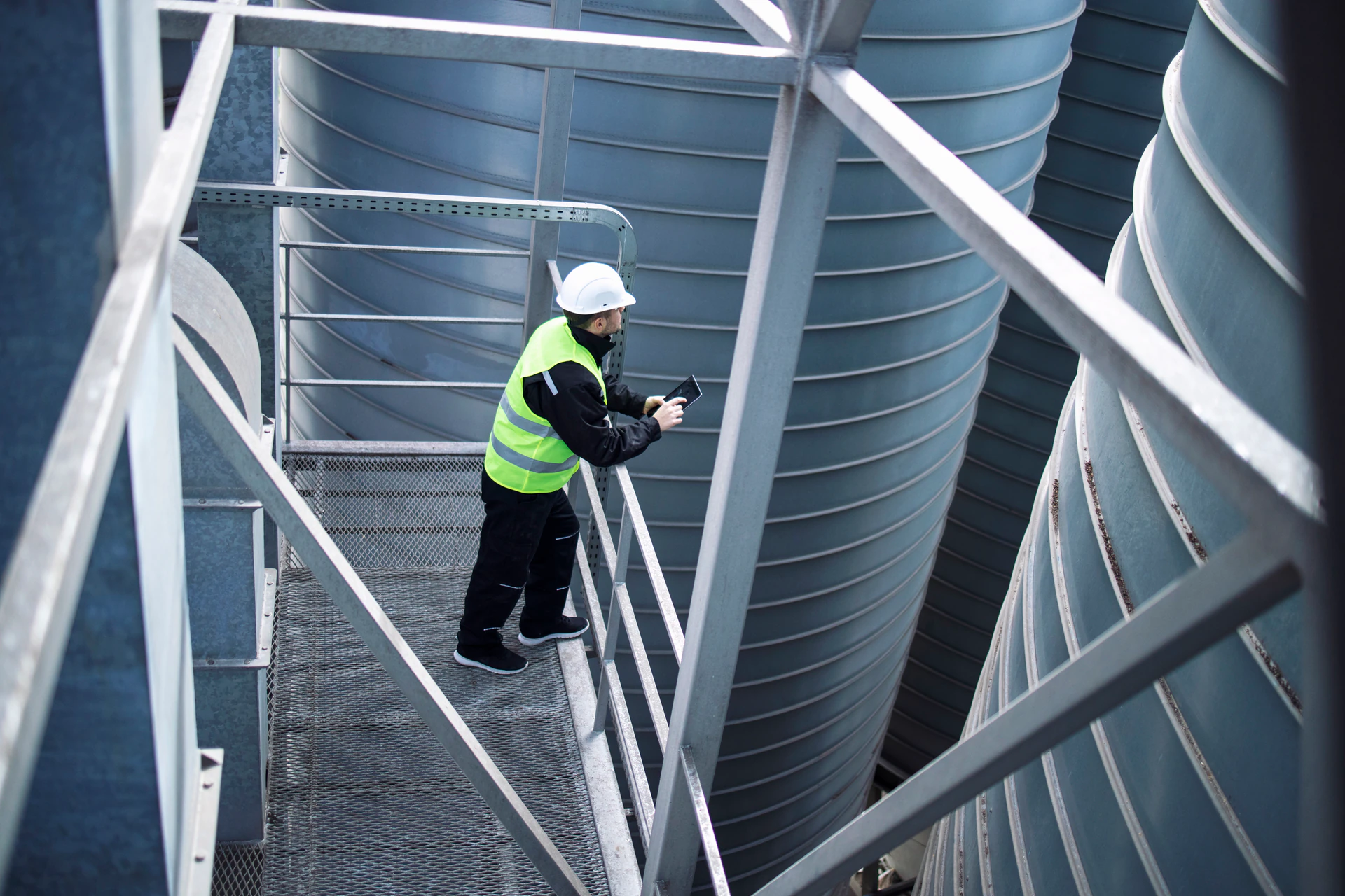 Trabalhador de silos de fabrica em pe na plataforma de metal entre tanques de armazenamento industrial e olhando um tablet sobre producao de alimentos